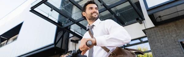 Visão de baixo ângulo de homem de negócios feliz no desgaste formal com saco de couro em pé perto de e-scooter, banner — Fotografia de Stock