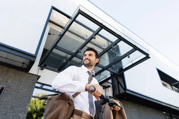 Vista de ángulo bajo de hombre de negocios feliz en desgaste formal con bolsa de cuero de pie cerca de e-scooter - foto de stock