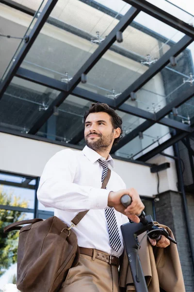 Низкий угол зрения веселого бизнесмена в формальной одежде с кожаной сумкой, стоящей рядом с электронным скутером — стоковое фото