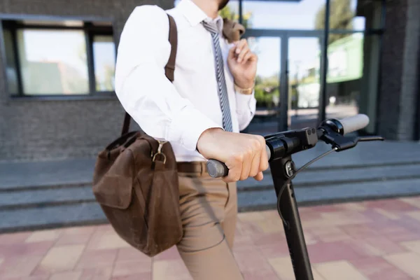 Vista recortada de hombre de negocios en ropa formal de pie cerca de e-scooter con bolso de cuero sobre fondo borroso - foto de stock