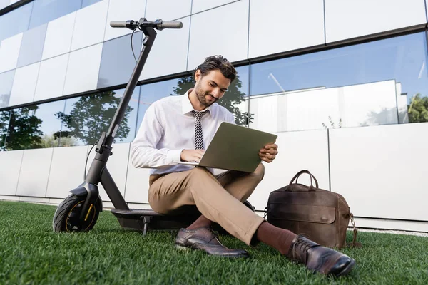 Homme d'affaires souriant en tenue formelle assis avec ordinateur portable près e-scooter sur l'herbe — Photo de stock