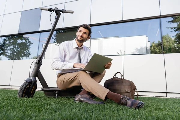 Freelance en tenue formelle assis avec ordinateur portable près e-scooter sur herbe — Photo de stock