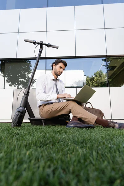 Businessman in formal wear sitting with laptop near e-scooter on grass — Stock Photo