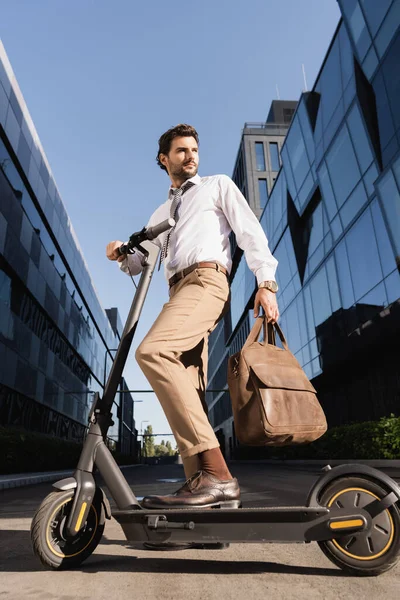 Visão de baixo ângulo de homem de negócios em desgaste formal segurando saco de couro e de pé perto de scooter elétrico — Fotografia de Stock