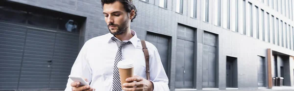 Бородатий бізнесмен в офіційному одязі тримає паперовий стаканчик і використовує смартфон, банер — стокове фото