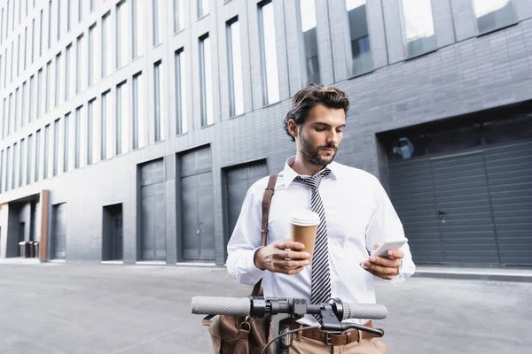 Bearded businessman in formal wear holding paper cup and using smartphone near e-scooter — Stock Photo