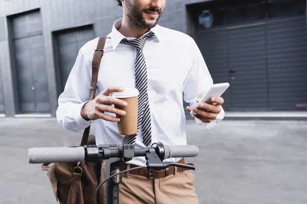 Обрезанный вид бородатого бизнесмена в формальной одежде, держащего бумажную чашку и использующего смартфон рядом с электронным скутером — стоковое фото
