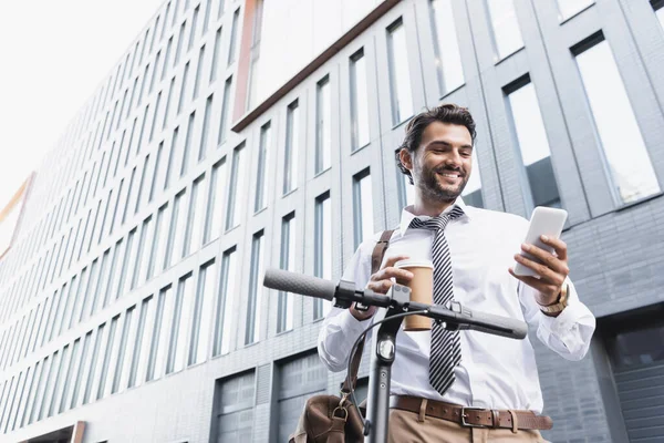 Vista de ángulo bajo de hombre de negocios feliz en el desgaste formal sosteniendo taza de papel y el uso de teléfono inteligente cerca de e-scooter - foto de stock