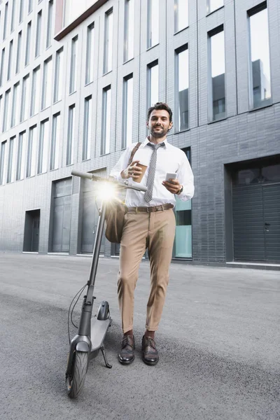 Full length of cheerful businessman in suit holding paper cup and using smartphone near e-scooter — Stock Photo