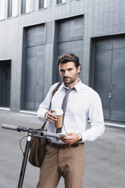 Homem de negócios barbudo em terno segurando café para ir e usando smartphone perto de e-scooter — Fotografia de Stock