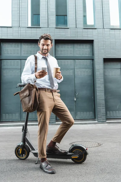 Повна довжина щасливого бізнесмена в офіційному носінні, що тримає паперовий стаканчик і смс на смартфоні поблизу електронного скутера — стокове фото