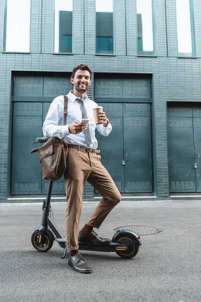 Piena lunghezza di uomo d'affari sorridente in abbigliamento formale in possesso di tazza di carta e sms su smartphone vicino e-scooter — Foto stock