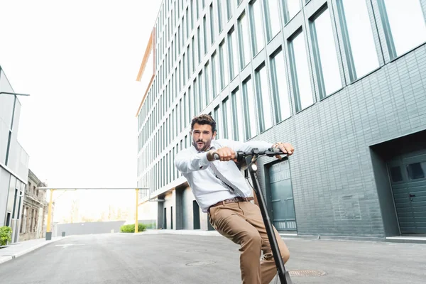 Empresario sorprendido en el desgaste formal montar scooter eléctrico cerca del edificio - foto de stock