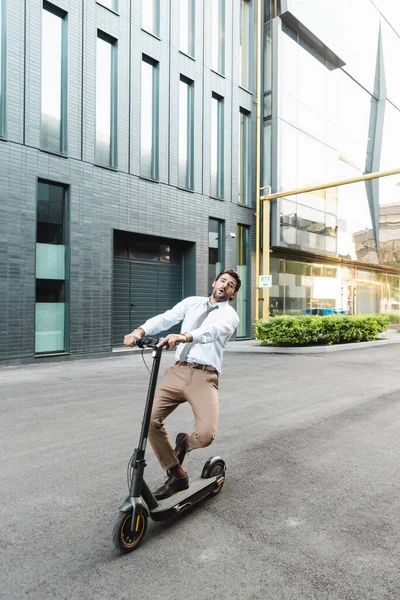 Homme d'affaires étonné en tenue formelle équitation scooter électrique près du bâtiment — Photo de stock