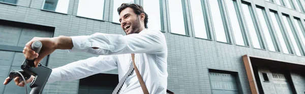 Vista del ángulo bajo del empresario sorprendido en el desgaste formal que monta la vespa eléctrica cerca del edificio, bandera - foto de stock