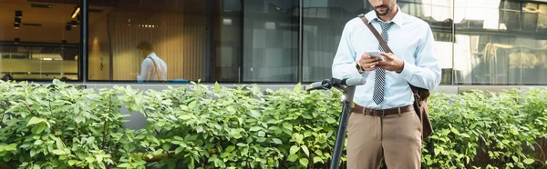 Vista cortada de homem de negócios mensagens de texto no smartphone enquanto estava perto de e-scooter, plantas e construção, banner — Fotografia de Stock