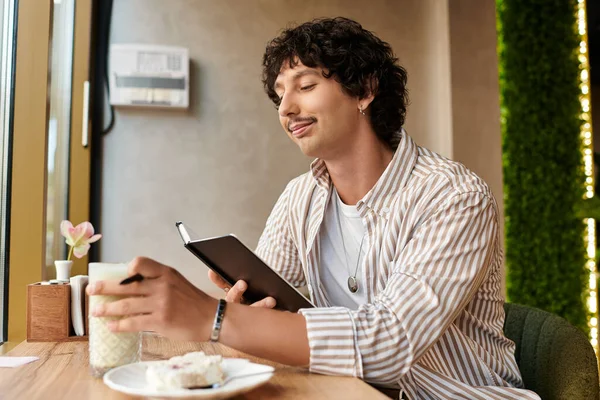 Ein gutaussehender junger Mann entspannt in einem Café, nippt an einem Drink und schreibt in sein Tagebuch. — Stockfoto