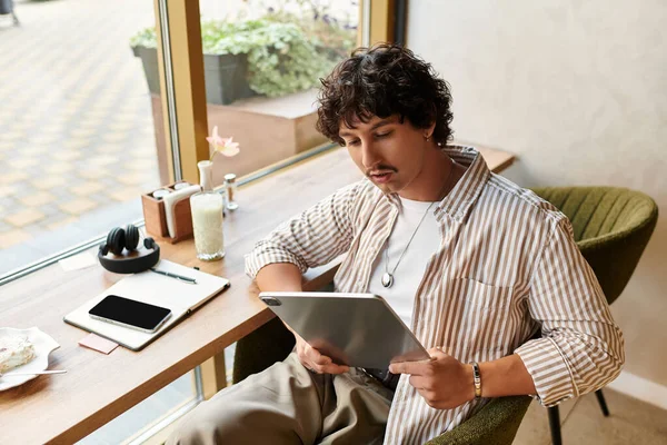 Schöner junger Mann sitzt an einem Cafétisch, in sein Tablet vertieft, inmitten einer entspannten Atmosphäre. — Stockfoto