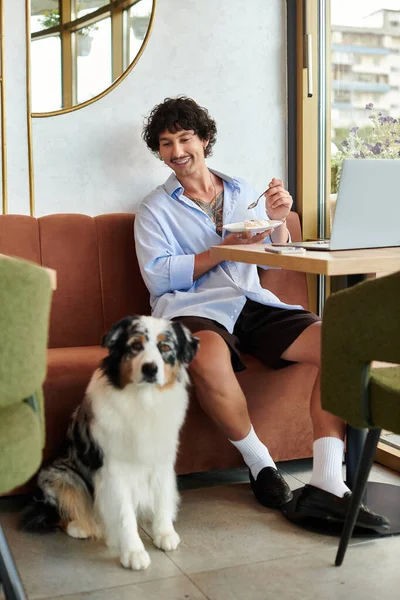 Um jovem com cabelo encaracolado desfruta de sua refeição em um café, enquanto seu cão relaxa nas proximidades. — Fotografia de Stock