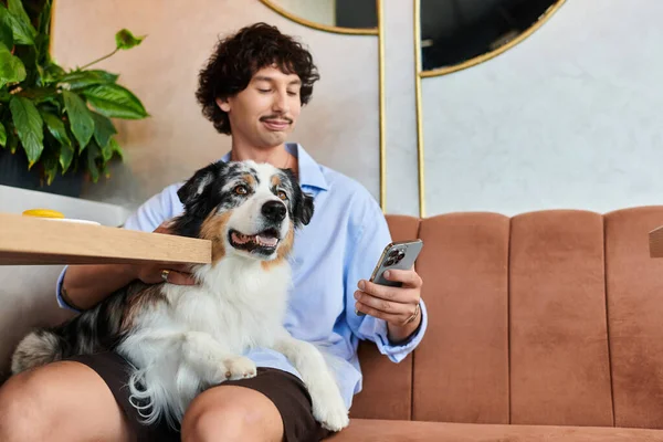 Ein junger Mann genießt die Zeit mit seinem Hund, während er in einem entzückenden Café sein Handy kontrolliert. — Stockfoto