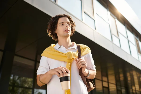 Красивый молодой человек в элегантной одежде прогуливается за пределами современной структуры, потягивая кофе. — стоковое фото