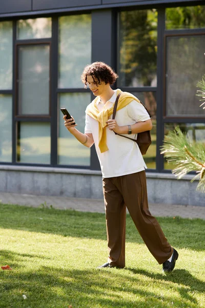 Handsome young man in stylish attire strolls on green grass, absorbed in the screen of his phone. — Stock Photo