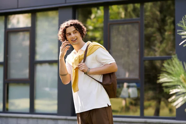 Ein junger Mann in schicker Freizeitkleidung lächelt und hält ein Telefon vor einem modernen Gebäude. — Stockfoto