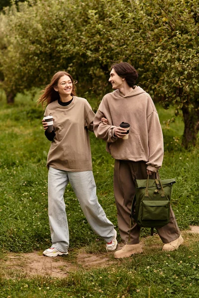 Любящая пара идет рука об руку по зеленому ландшафту, делясь улыбками и кофе. — стоковое фото