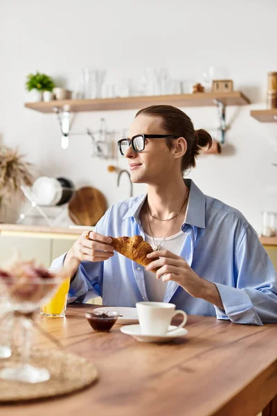 Красивый, женственный молодой человек улыбается, наслаждаясь круассаном и кофе за своим кухонным столом. — стоковое фото