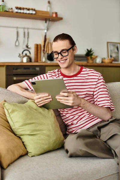 Ein jugendlicher, gutaussehender Mann sitzt gemütlich auf einer Couch und lächelt, während er sein Tablet bedient. — Stockfoto