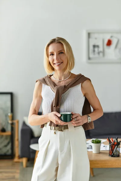 Una hermosa mujer está de pie en su acogedora casa, sonriendo mientras sostiene una taza de café, irradiando calor. - foto de stock