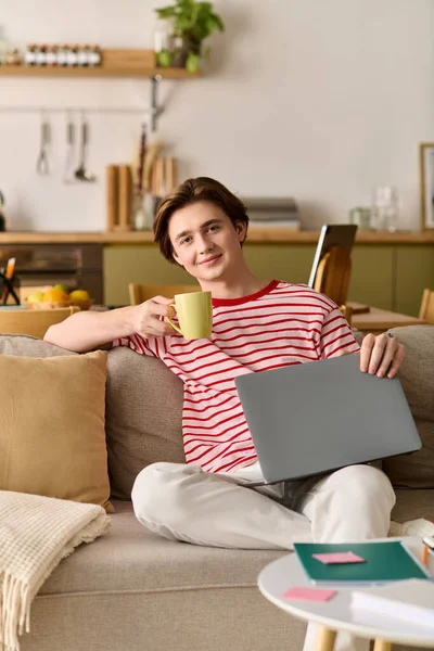 Um jovem estuda online enquanto relaxa em casa, sorvendo de uma caneca e sorrindo. — Fotografia de Stock
