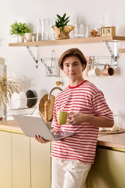 Молодой человек учится дома, используя ноутбук для доступа к электронному обучению, потягивая кофе. — стоковое фото