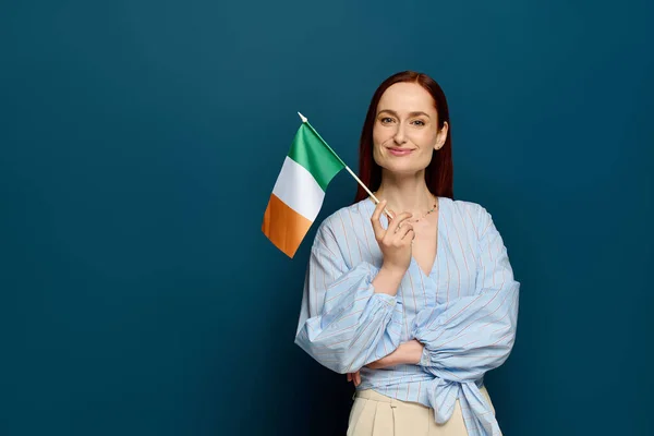 A language teacher smiles confidently while holding an Irish flag against a blue background. - foto de stock