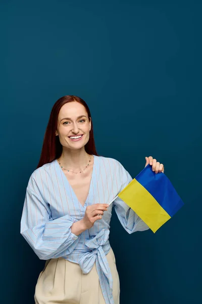 A language teacher with red hair joyfully holds a flag against a blue background in her studio. — стоковое фото
