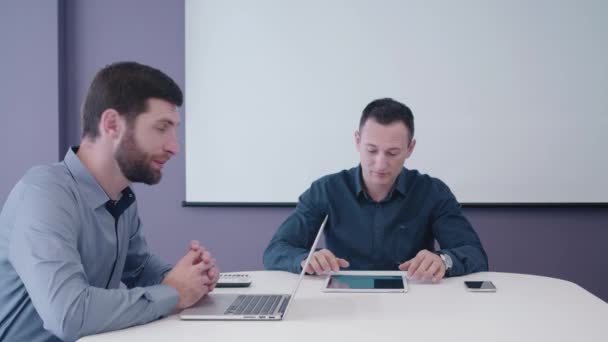 Dois empresários de meia-idade sentados na sala de reuniões com tablet e laptop na mesa e discutindo educadamente a ideia de inicialização com seu interlocutor atrás da câmera — Vídeo de Stock