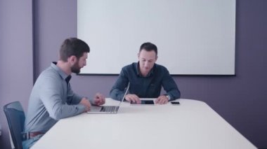 iki, tıraş ve sakallı, orta yaşlı işadamları tablet ve masanın üstünde laptop ve gülümseyen ve nazikçe fikirlerini tartışıyor jestleri ile Toplantı odasında otururken ateş. 4 k Orta