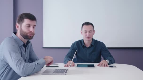 Dois empresários caucasianos de meia-idade sentados com tablet e laptop na mesa na sala de reuniões debatendo e discordando com seu interlocutor atrás da câmera — Vídeo de Stock