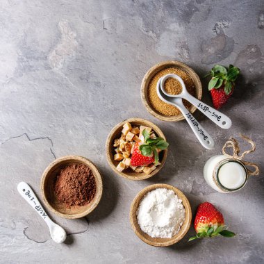 Çikolata kupa kek yemek pişirmek için malzemeler. Un, kakao tozu, şeker, karamel ahşap kase, süt, çilek kaşıklarla gri doku arka plan üzerinde görev yaptı. Üstten görünüm alanı. Kare görüntü