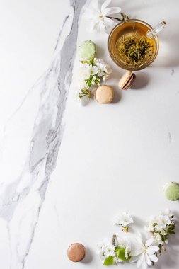Cam fincan yeşil çay Fransız tatlı macaroons, bahar çiçekleri beyaz Manolya ve beyaz mermer doku arka plan üzerinde kiraz çiçek açan dalları ile. Üstten Görünüm, kopya alanı.