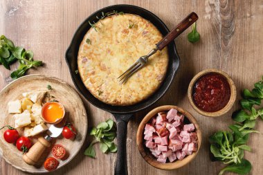 İspanyol patates omlet tortilla pastırma ile sos ve malzemeler yukarıda ile dökme demir tavada ahşap arka plan üzerinde servis. Üstten Görünüm, uzay.
