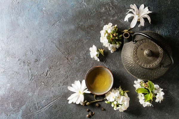 传统陶瓷杯的热绿茶与黑铁茶壶 春花白玉兰和樱桃盛开的树枝在深蓝色的纹理背景下 顶部视图 复制空间 — 图库照片