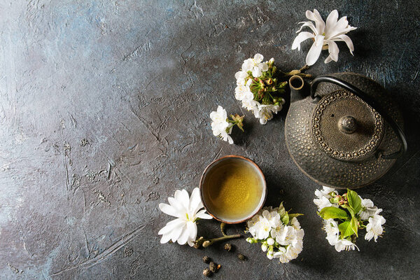 Traditional ceramic cup of hot green tea with black iron teapot, spring flowers white magnolia and cherry blooming branches over dark blue texture background. Top view, copy space.