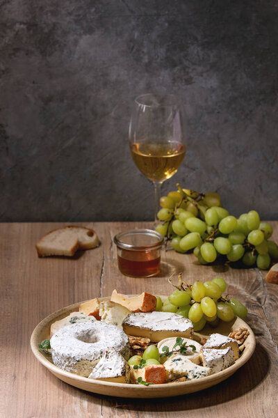 Cheese plate assortment of french cheese served with honey, walnuts, bread and grapes on ceramic plate and glass of white wine over wooden table with grey wall as background.