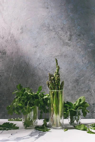 生調理有機グリーン アスパラガス コリアンダー ハーブと灰色のテクスチャの壁の背景としての白いキッチン テーブルの上のガラスの瓶で新鮮なミントの束 コピー スペース — ストック写真