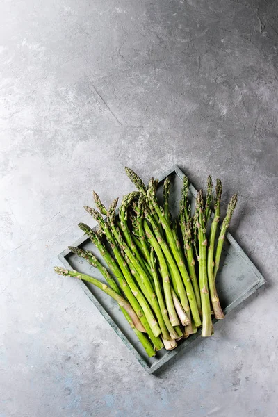 Çiğ çiğ organik yeşil kuşkonmaz içinde gri doku arka plan üzerinde ahşap tepsi. Üstten Görünüm, kopya alanı.