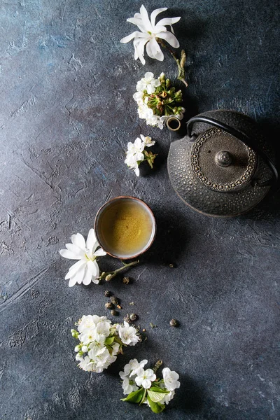 传统陶瓷杯的热绿茶与黑铁茶壶 春花白玉兰和樱桃盛开的树枝在深蓝色的纹理背景下 顶部视图 复制空间 — 图库照片