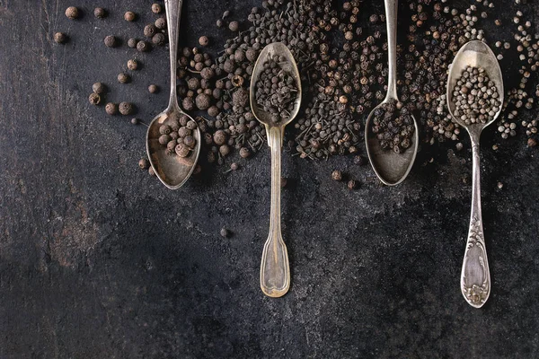 Farklı siyah biber yenibahar, yenibahar, rahipler biber, karabiber eski siyah demir doku arka plan üzerinde vintage kaşık içinde çeşididir. Üstten görünüm, uzay.