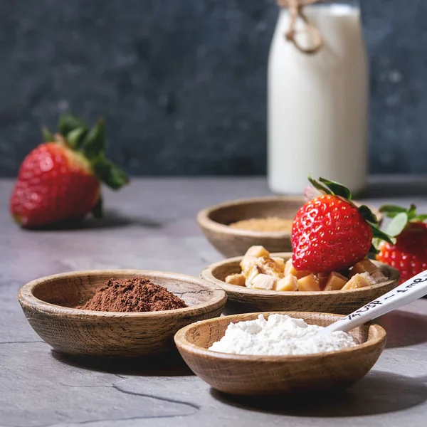 Ingredients for cooking chocolate mug cakes. Flour, cocoa powder, sugar, caramel in wooden bowls, milk, strawberries and mint served with spoons and mug over grey kitchen table. Square image