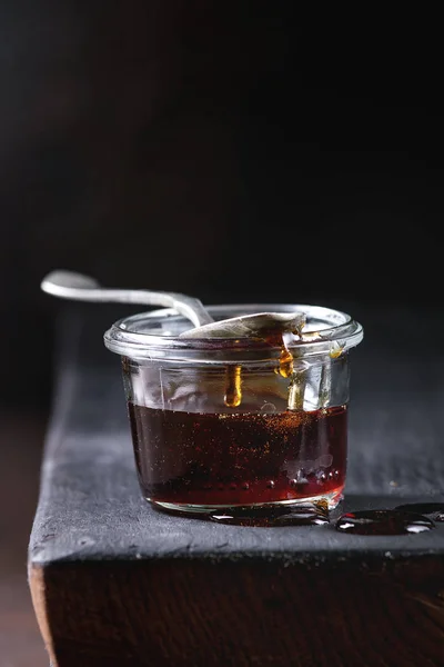 Homemade liquid transparent brown sugar caramel in glass jar standing on black wooden board with spoon. Close up